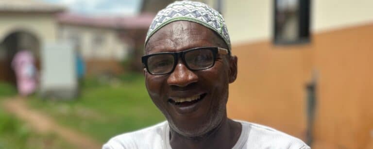 A smiling man wearing eyeglasses, a white t-shirt, and a patterned cap poses outdoors with arms crossed.