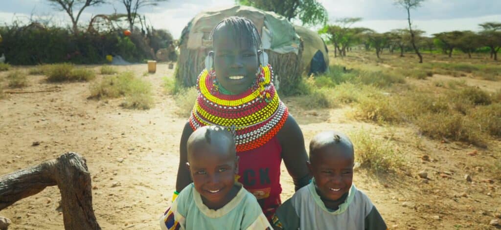 Ethekon Loote wearing traditional colorful beaded neckwear smiles alongside her two young children outdoors in Samburu, Kenya.