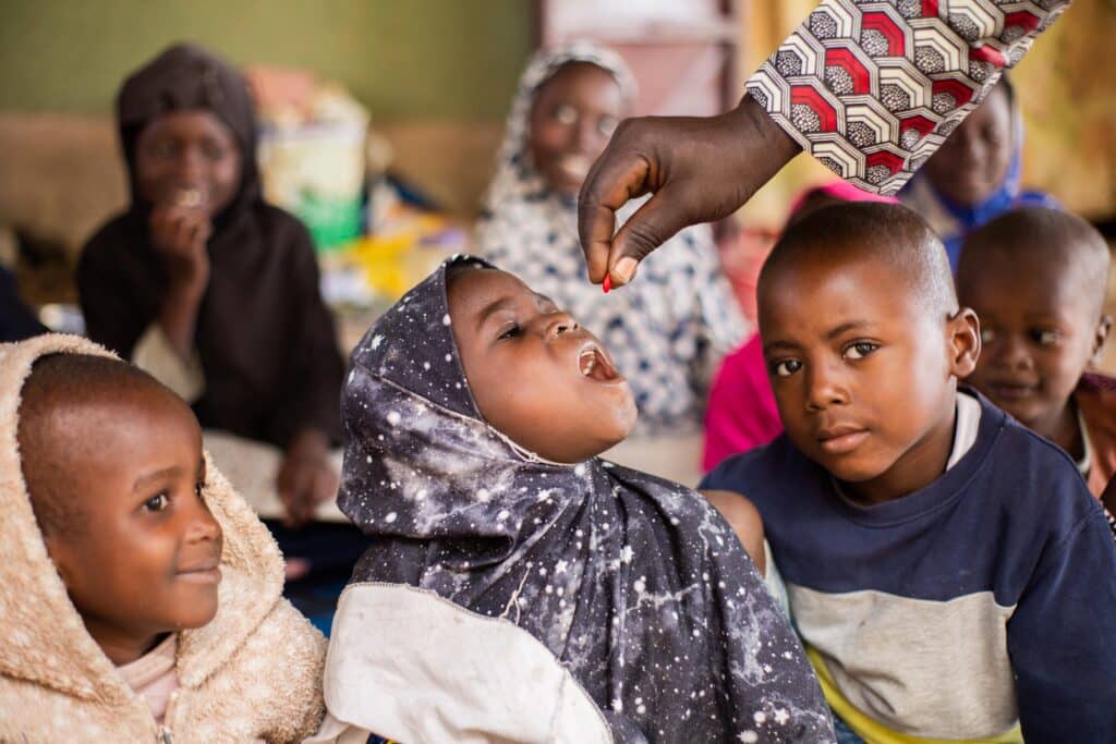 A healthcare worker administers a small red vitamin A capsule to a young girl wearing a starry patterned headscarf in Garoua, Cameroon, while other children look on.