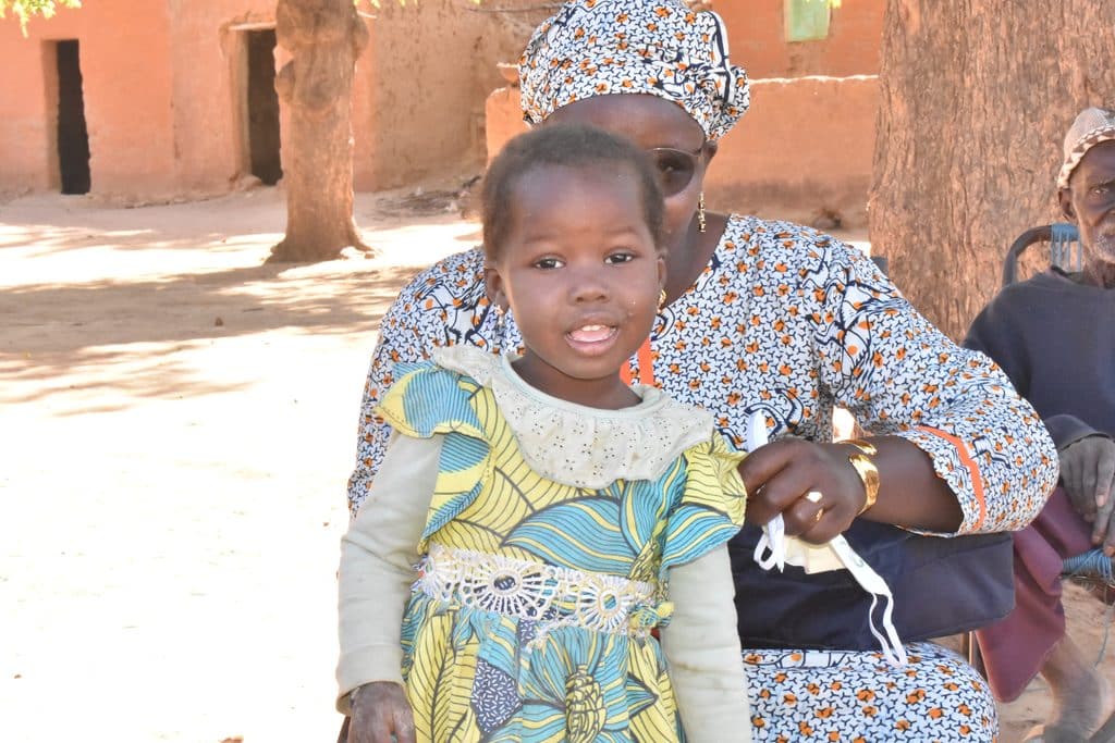 A young girl in a yellow dress smiling standing in front of her grandmother holding a face mask during a health campaign in Banatomo, Mali.