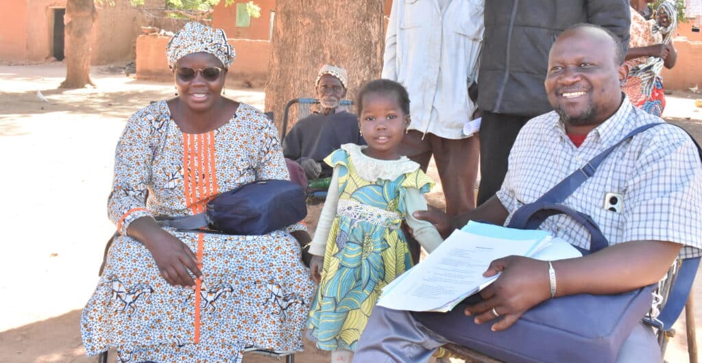 Five-year-old Maimouna poses with her grandmother and health workers outdoors under a large tree.