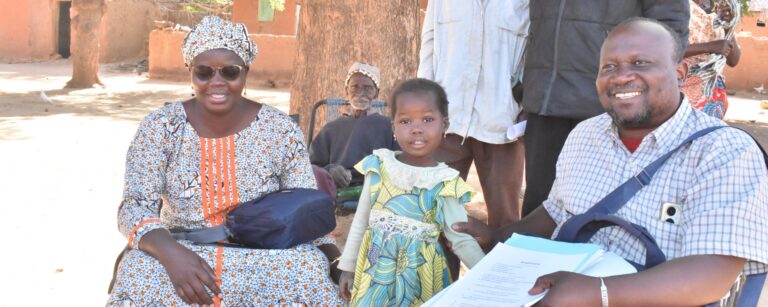 Five-year-old Maimouna poses with her grandmother and health workers outdoors under a large tree.
