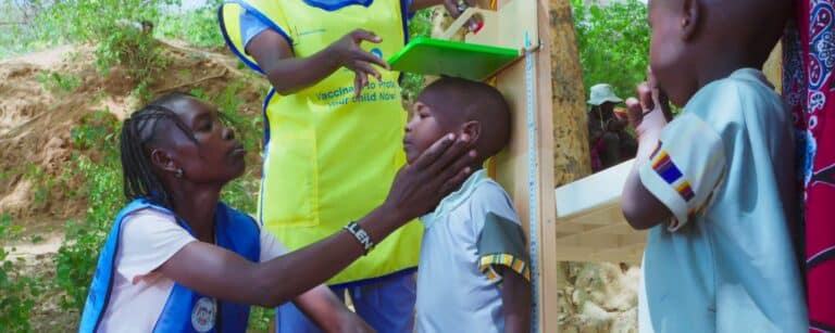 Two health workers in bright yellow and blue vests gently measure a young child's height using a wooden stadiometer outdoors in a rural setting, while another child looks on.