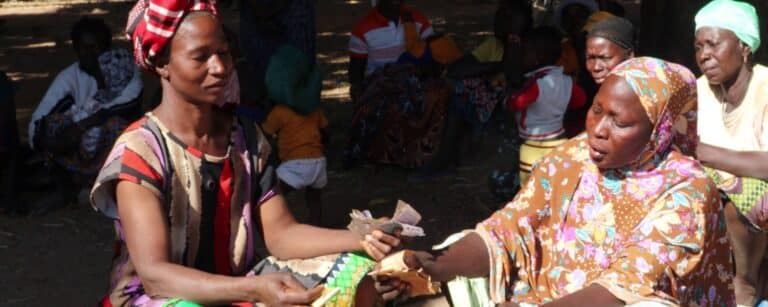 Two women wearing headscarves and colourful patterns sit facing each other outdoors during a community gathering, exchanging banknotes directly hand-to-hand while other community members look on in the background.