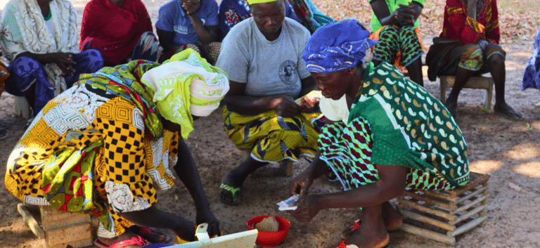 A group of women in colorful headscarves gathered outdoors in a circle during a Village Savings and Loan Association (VSLA) meeting, counting money and keeping records.